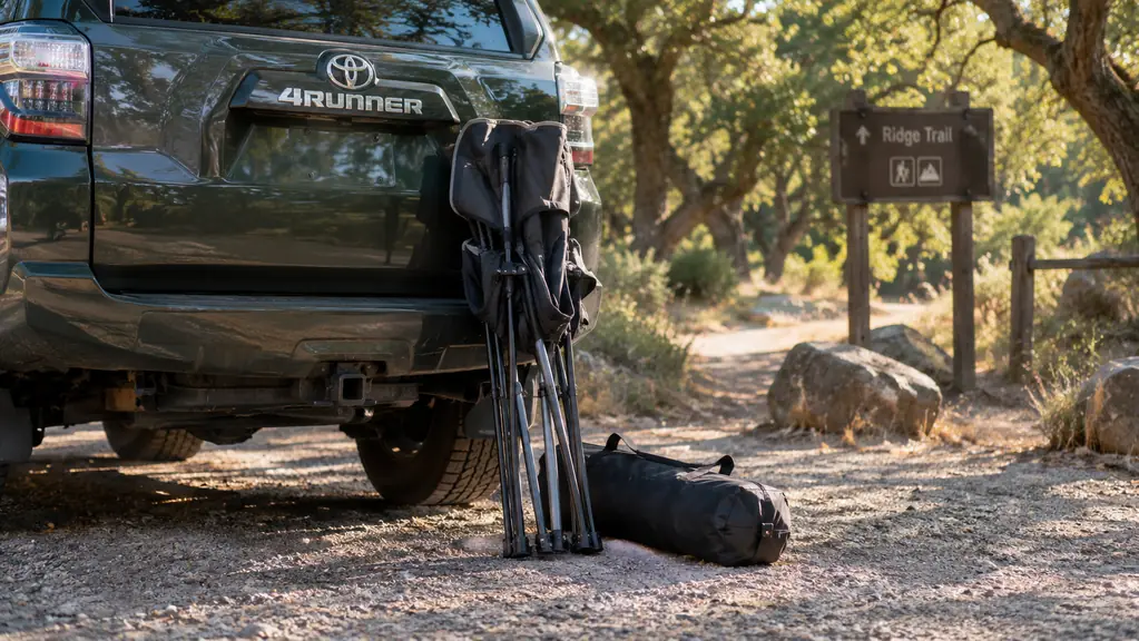 A folded camp chair and its carry bag leaning against the tailgate of a vehicle at a park trailhead in the morning