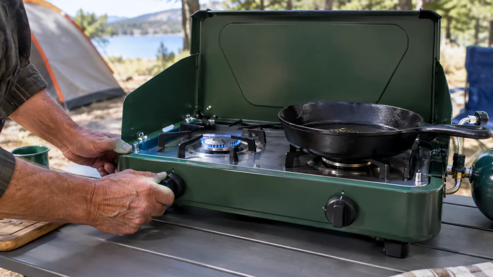 Hands turning the knob to light a two-burner propane stove on a camp table, with a skillet on the other burner.