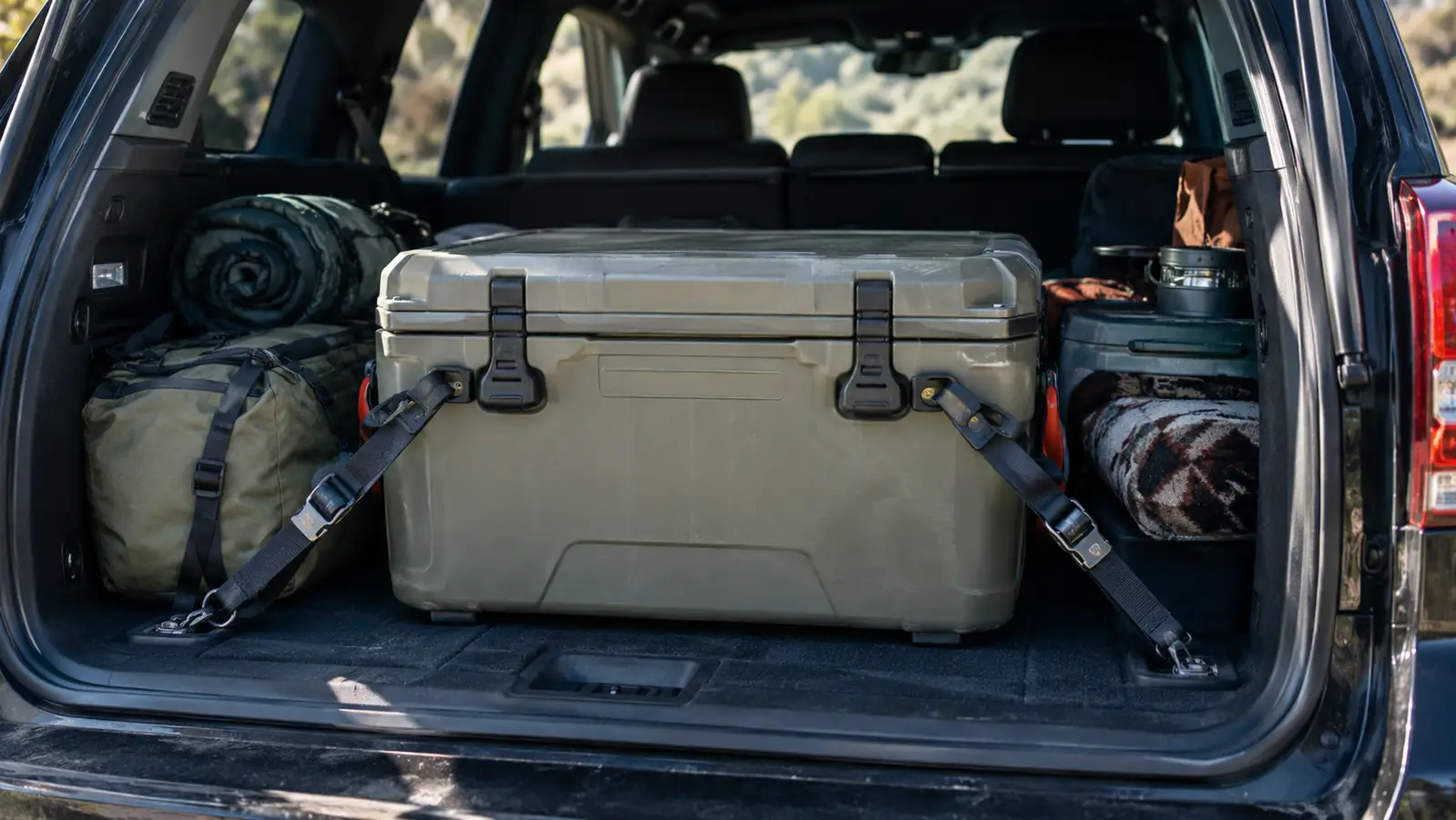 A chest-style cooler secured in the back of an SUV with tie-down straps, camping gear packed around it